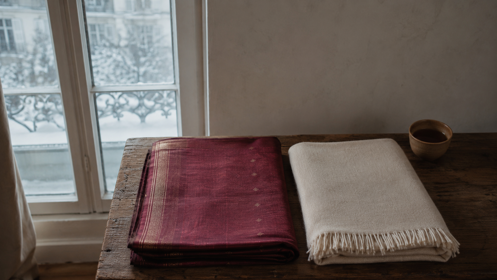 A maroon silk saree folded beside a cream wool shawl on a wooden bench in Munich winter light