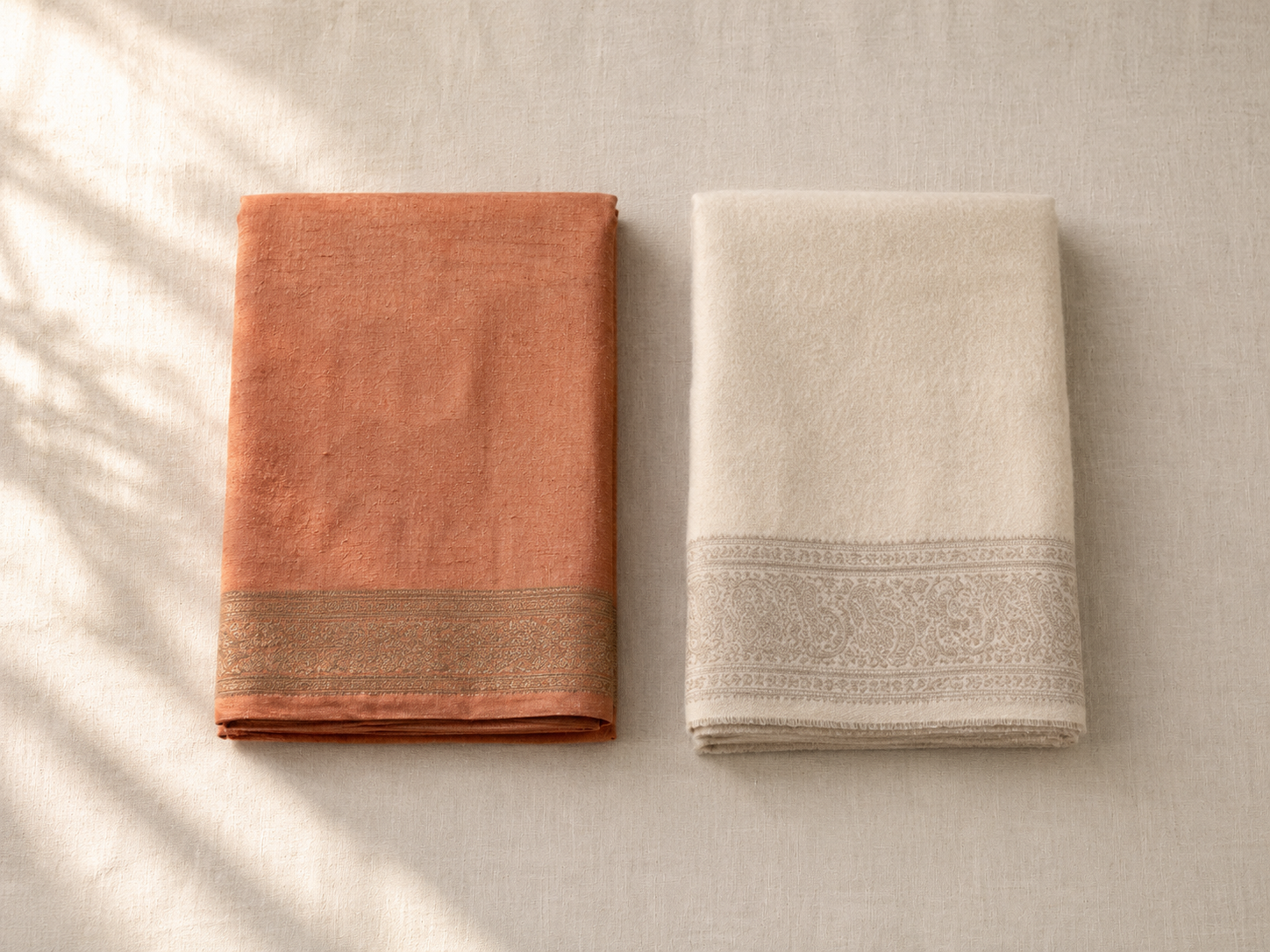 A folded warm-toned tussar silk saree beside a cream pashmina wool saree on a wooden surface