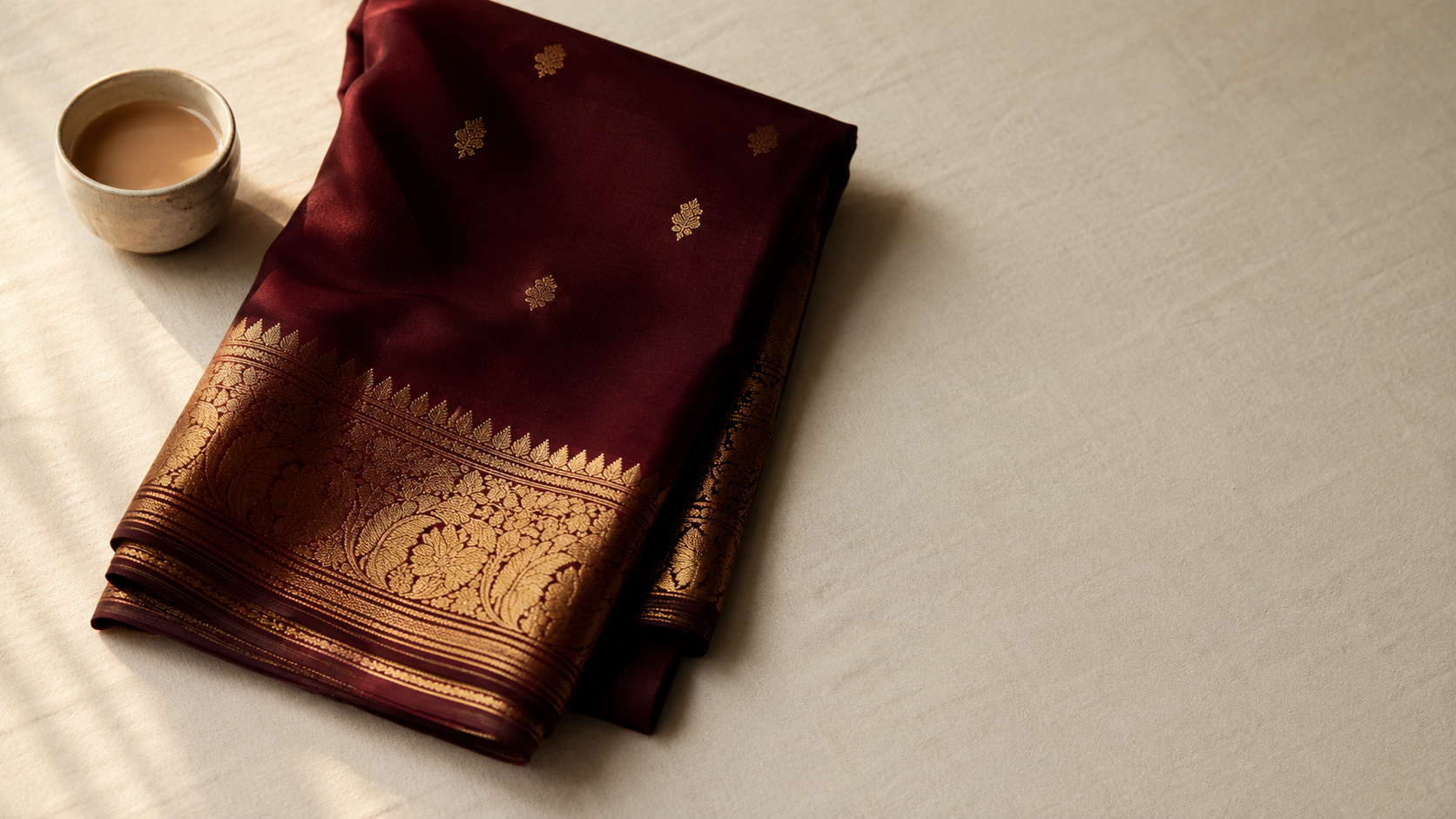 A folded maroon silk saree with gold zari border on cream linen — IndiaNiva guide to what is a saree